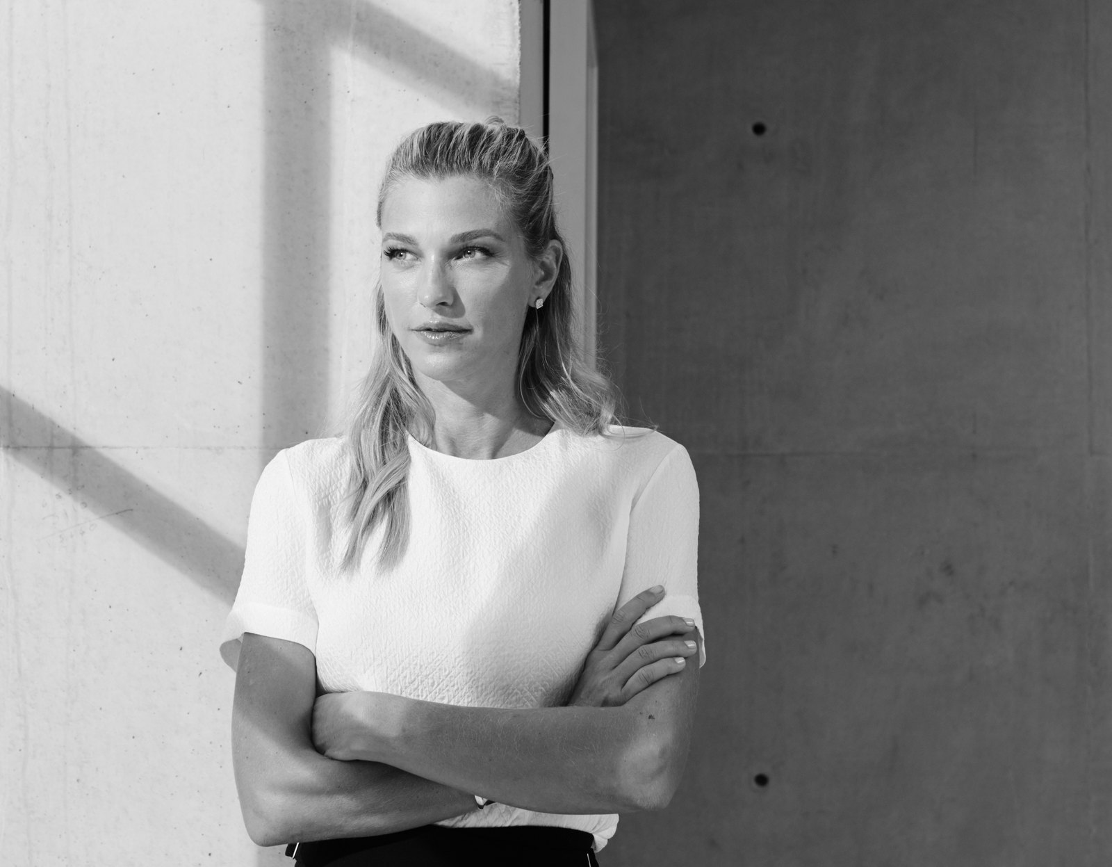 Ritratto in bianco e nero di Nicole Junkerman appoggiata a una parete di cemento, con le braccia conserte e lo sguardo rivolto verso la luce